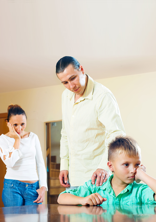 sad young boy sat at table with parents stood behind trying to provide comfort